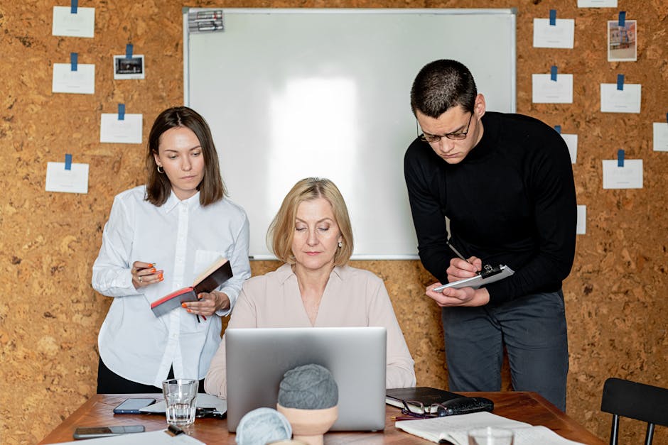 A diverse team collaborates in an office meeting, using a laptop and taking notes.